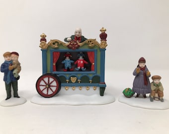 Puede incluir: Un conjunto de figuritas de porcelana que representa un espectáculo de títeres con un carro verde y azul, un titiritero y dos títeres. Dos niños miran el espectáculo, mientras que otro niño se para con una cesta de bolas verdes.