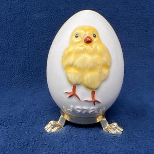 Puede incluir: Un huevo de cerámica blanca con un diseño de pollito amarillo y patas rojas. El huevo está sobre un soporte dorado con tres patas. El soporte tiene el año 1976 inscrito en él.