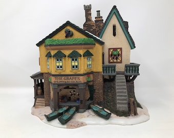 Puede incluir: Un edificio de cerámica en miniatura con una fachada amarilla y un techo verde. El edificio tiene un letrero que dice "The Grapes Old, Mild & Bitter Ales". Hay pequeños botes frente al edificio.