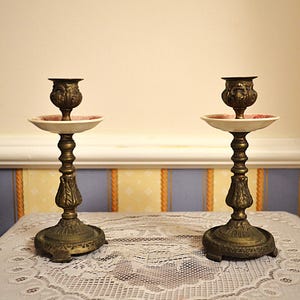 May include: A pair of matching antique-style brass candlesticks featuring ornate, textured stems and circular bases. Each candlestick includes a white, shallow, saucer-like drip tray positioned beneath the candle holder cup.