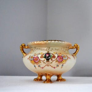 May include: A vintage cream-coloured porcelain bowl with gold detailing. The bowl has floral patterns in pink, yellow, and blue, with gold handles and feet. The rim has a scalloped edge, and the design is ornate and decorative, suitable for display.