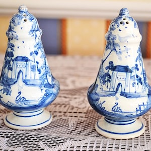 Op de afbeelding: Een paar zout- en peperstrooiers van wit keramiek met een handgeschilderd blauw landschapsontwerp. Elke strooier heeft een klokvormig lichaam, een ronde voet en een koepelvormige bovenkant met kleine gaatjes voor het doseren van kruiden.