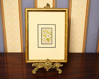 Op de afbeelding: Een gouden lijst met een bloemenprint van witte bloemen met gele centra. De lijst staat op een decoratief, goudkleurig ezel, met een donker houten oppervlak en behang met patronen op de achtergrond.