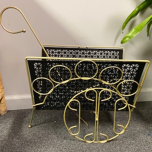 May include: Gold metal magazine rack with a black wire mesh design. The rack has a decorative, circular design on the bottom and a hook on the top.