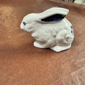 May include: A ceramic rabbit figurine with a crackle glaze finish. The rabbit has blue accents on its ears, eyes, and a floral design on its side. The figurine is a light beige color.
