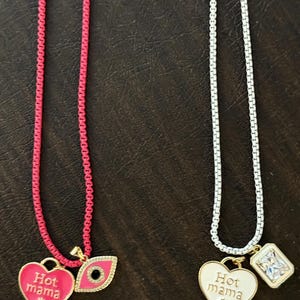 May include: Two gold necklaces with charms. The necklace on the left has a pink chain and a pink heart charm with the text "Hot mama" and a blue evil eye charm. The necklace on the right has a silver chain and a white heart charm with the text "Hot mama" and a clear square charm.
