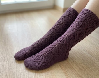 May include: A pair of mid-calf length knitted socks in a deep plum purple color. The socks feature a textured, repeating diamond-shaped cable knit pattern that extends from the ribbed cuffs down to the rounded toe area of the foot.