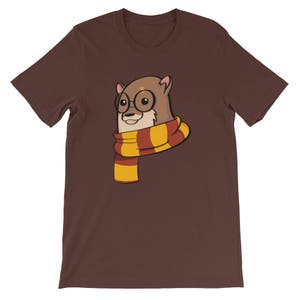 May include: A brown t-shirt featuring a cartoon otter wearing round glasses and a scarf with red and yellow stripes. The otter has a lightning bolt on its head. The shirt is a solid color.