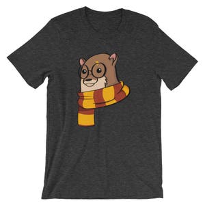 May include: Dark gray t-shirt featuring a cartoon otter wearing round glasses and a scarf with red and yellow stripes. The otter has a lightning bolt on its forehead. The shirt is made of a soft material.