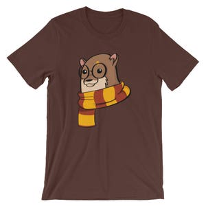 May include: Brown t-shirt featuring a cartoon otter wearing round glasses and a striped scarf in shades of red, yellow, and gold. The otter has a lightning bolt scar on its forehead. The shirt is a solid brown color.