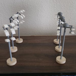 May include: Several miniature, abstract sculptures with gray stems and white, rectangular tops are arranged on a dark wooden surface. Each sculpture has a circular, beige base. The background includes a shelf with video games.