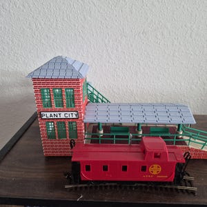 May include: A model train set featuring a red caboose and a brick station with the text "PLANT CITY." The station has a gray roof and green accents. The caboose is on a track.