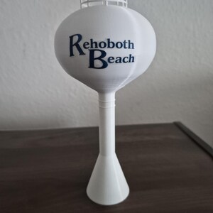 Könnte beinhalten: Ein weißes 3D-gedrucktes Wasserturm-Modell mit einem kugelförmigen Tank, einem Geländer an der Oberseite und einem konischen Sockel. Der Text Rehoboth Beach ist in dunkelblauen Buchstaben auf der Vorderseite des kugelförmigen Tanks aufgedruckt.