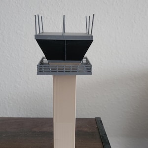 Könnte beinhalten: Ein graues und beiges Modell eines Flughafenturm. Der Turm hat einen quadratischen Sockel und ein schwarzes, abgewinkeltes Dach mit einem Geländer um die Spitze.