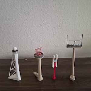Könnte beinhalten: Vier Miniatur-Architekturmodelle von Kommunikationstürmen und -strukturen. Die Modelle sind weiß, beige und rot und sind auf einer dunkelbraunen Oberfläche angeordnet. Die Modelle umfassen einen Turm mit einer Kugel oben, einen Turm mit roten Antennen und einen Turm mit rechteckiger Oberseite.