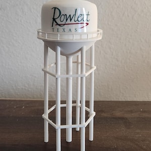May include: A white miniature water tower with the text "Rowlett TEXAS" in black and red. The model has a cylindrical top and a lattice-like support structure. It is displayed on a dark brown surface.