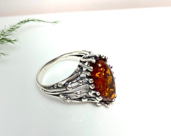 May include: A silver ring with a large amber gemstone set in a detailed, intricate design. The ring has a wide band and the gemstone is surrounded by delicate silver branches.