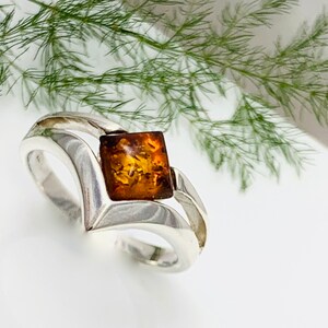 Peut inclure: Une bague en argent avec une pierre précieuse ambrée carrée sertie au centre. La bague a un design unique et moderne avec une bande incurvée.