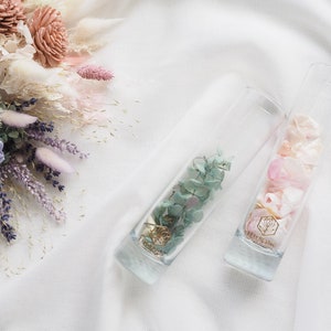 May include: Two clear glass vases filled with dried flowers. The vase on the left contains light green dried flowers, while the vase on the right contains pink dried flowers. Both vases have a gold label with a floral design.