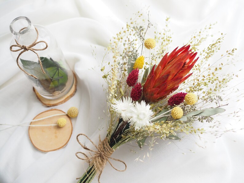 May include: A dried flower bouquet with a red protea, white billy buttons, yellow craspedia, and dried grasses tied with twine.