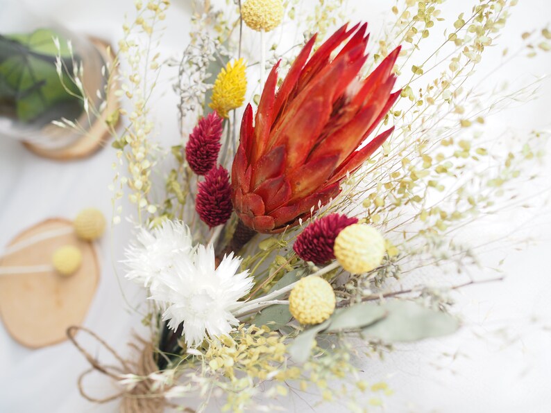 May include: A dried flower bouquet with a red protea, white billy buttons, burgundy statice, and yellow craspedia.