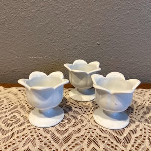 May include: A set of three white ceramic egg cups, each shaped like an open flower blossom with six rounded petals, a smooth curved bowl, and a circular pedestal base, arranged in a cluster on a flat surface.