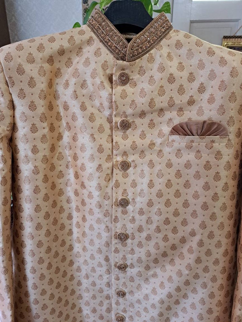 May include: A beige kurta with a brown patterned design and a brown velvet pocket. The kurta has a high collar with a beaded trim and a button closure.