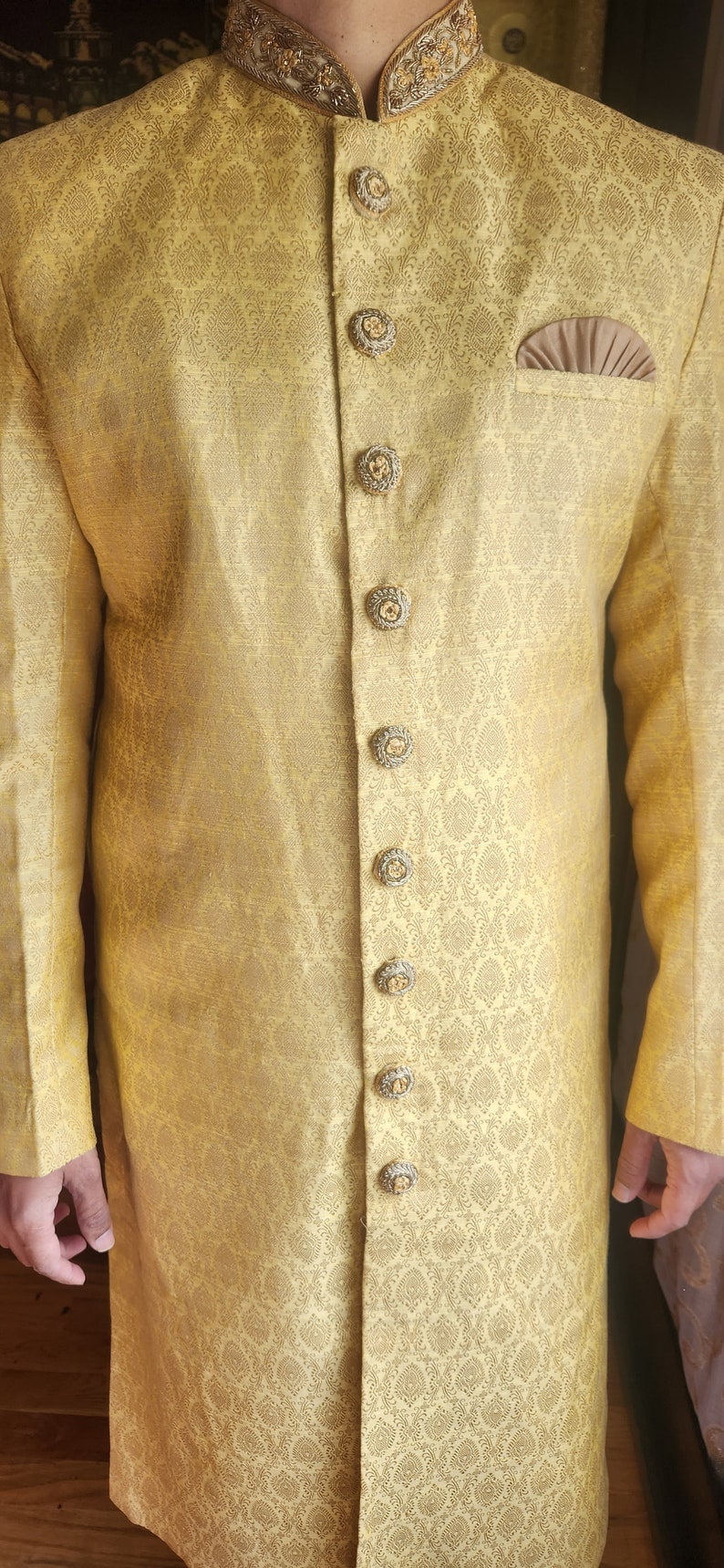 K&ouml;nnte beinhalten: Goldgelbe Jacke mit langen &Auml;rmeln und aufwendigen, sich wiederholenden Mustern. Die Jacke hat dekorative Kn&ouml;pfe und eine kleine Tasche. Kragen und Kn&ouml;pfe sind mit Gold verziert. Es ist ein formelles Kleidungsst&uuml;ck.