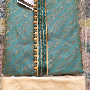 May include: Teal and gold embroidered kurta pajama set. The top has a mandarin collar, decorative buttons, and the brand name "I.ELEGANT". The set includes matching beige pants. The fabric has a subtle floral pattern.