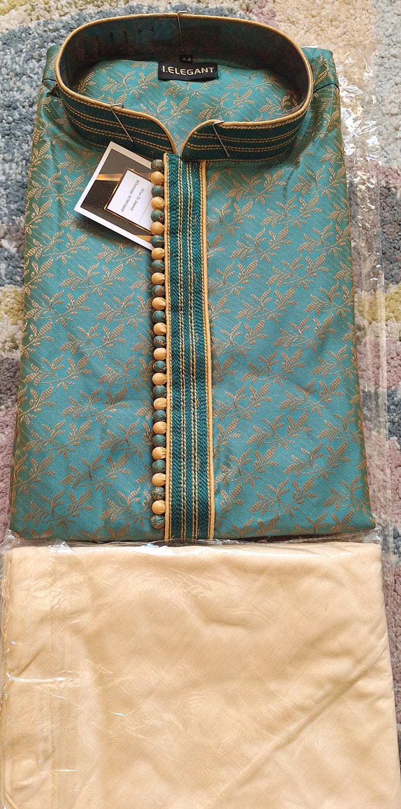 May include: Teal and gold embroidered kurta pajama set. The top has a mandarin collar, decorative buttons, and the brand name "I.ELEGANT". The set includes matching beige pants. The fabric has a subtle floral pattern.