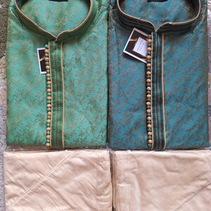 May include: Two silk kurta pajama sets. One is teal, the other is green, both with gold floral patterns. Each kurta has a mandarin collar and wooden bead accents. The matching pajama pants are a light beige color.