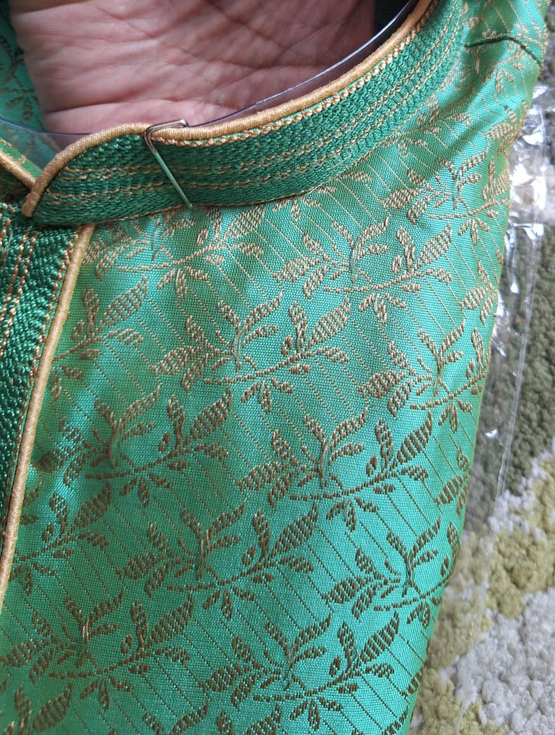 May include: Close-up of a green garment with gold floral embroidery. The collar and trim are a darker green with gold accents. The fabric has a subtle sheen, suggesting a silk or satin material.