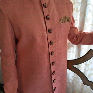 Puede incluir: Chaqueta de color coral con cuello mandar&iacute;n y botones florales ornamentados. La chaqueta presenta un patr&oacute;n sutil y repetitivo y un cuello bordado en contraste rojo oscuro y dorado. Se ve un pa&ntilde;uelo de bolsillo.