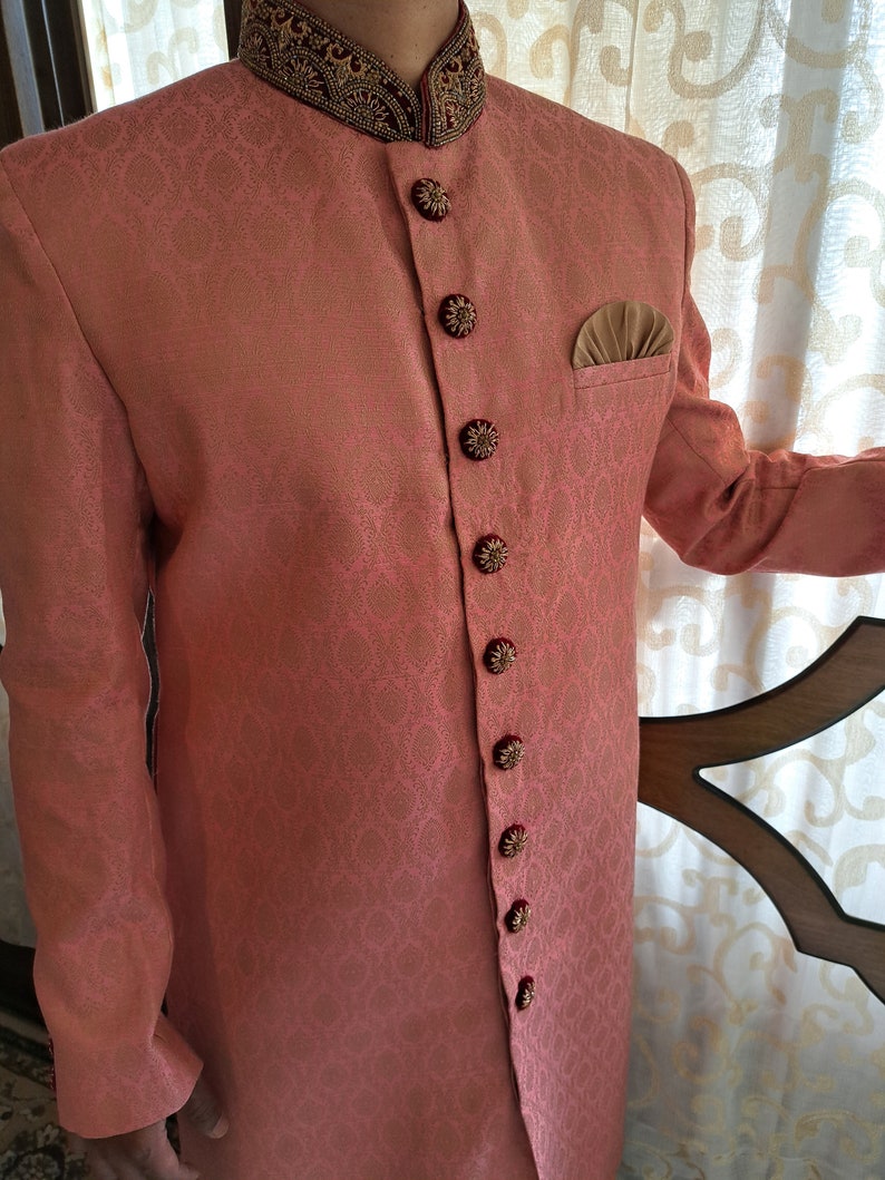 Puede incluir: Chaqueta de color coral con cuello mandar&iacute;n y botones florales ornamentados. La chaqueta presenta un patr&oacute;n sutil y repetitivo y un cuello bordado en contraste rojo oscuro y dorado. Se ve un pa&ntilde;uelo de bolsillo.