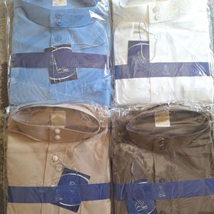 May include: Four different colored shirts in plastic packaging. The shirts are light blue, white, beige, and brown. Each shirt has a button-up collar and two buttons on the front. The shirts have a label with a bird logo and the text "Al-Salam" on them.