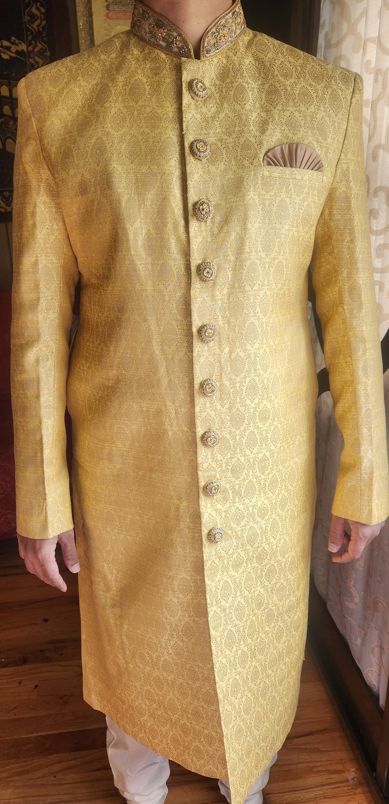 K&ouml;nnte beinhalten: Goldgelbe, lang&auml;rmlige Jacke mit Muster, verzierten Kn&ouml;pfen und Stehkragen. Die Jacke hat ein Einstecktuch und ist Teil eines formellen Outfits. Der Stoff hat ein subtiles, sich wiederholendes Design.