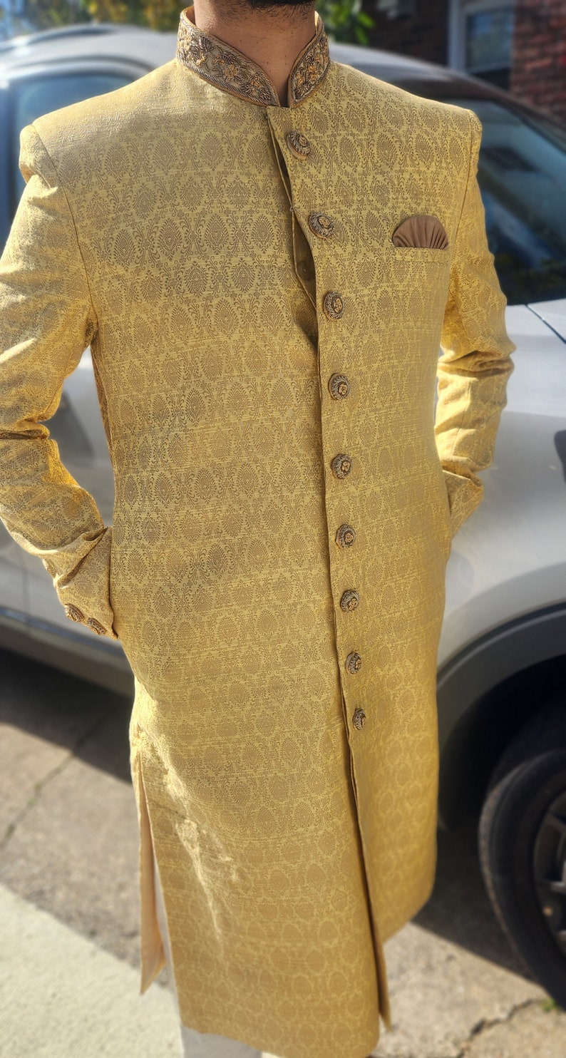 K&ouml;nnte beinhalten: Goldfarbene Jacke mit langen &Auml;rmeln und einem komplizierten, sich wiederholenden Muster. Die Jacke hat dekorative Kn&ouml;pfe, eine Tasche und einen detaillierten Kragen. Das Kleidungsst&uuml;ck ist Teil eines formellen Ensembles.