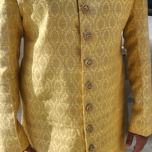 K&ouml;nnte beinhalten: Eine goldgelbe Jacke mit langen &Auml;rmeln und einem komplizierten Paisley-Muster. Die Jacke hat verzierte, goldfarbene Kn&ouml;pfe und einen dekorativen Kragen. Ein Einstecktuch ist sichtbar. Das Kleidungsst&uuml;ck ist Teil eines traditionellen formellen Outfits.