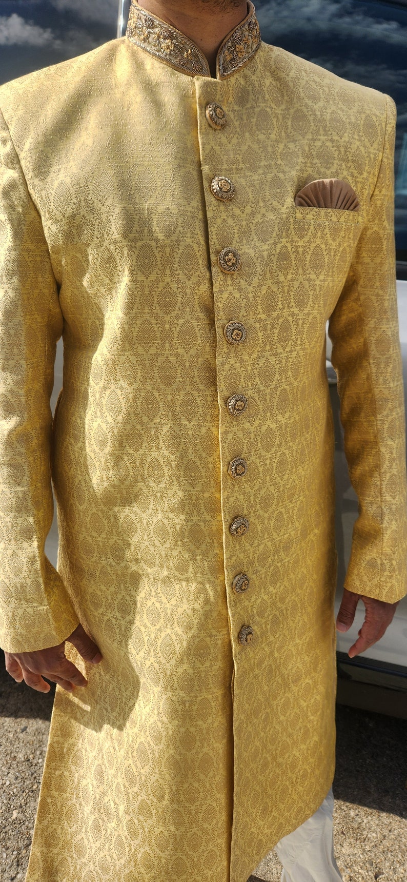 K&ouml;nnte beinhalten: Eine goldgelbe Jacke mit langen &Auml;rmeln und einem komplizierten Paisley-Muster. Die Jacke hat verzierte, goldfarbene Kn&ouml;pfe und einen dekorativen Kragen. Ein Einstecktuch ist sichtbar. Das Kleidungsst&uuml;ck ist Teil eines traditionellen formellen Outfits.