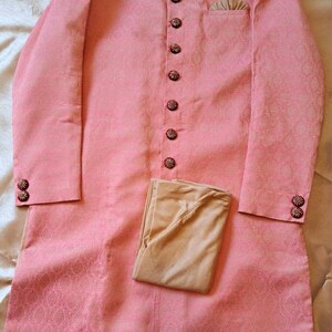 Puede incluir: Una t&uacute;nica rosa claro de manga larga con cuello mandar&iacute;n y botones decorativos. La t&uacute;nica tiene un sutil estampado floral y se combina con pantalones beige. El cuello y los pu&ntilde;os presentan bordados ornamentados.