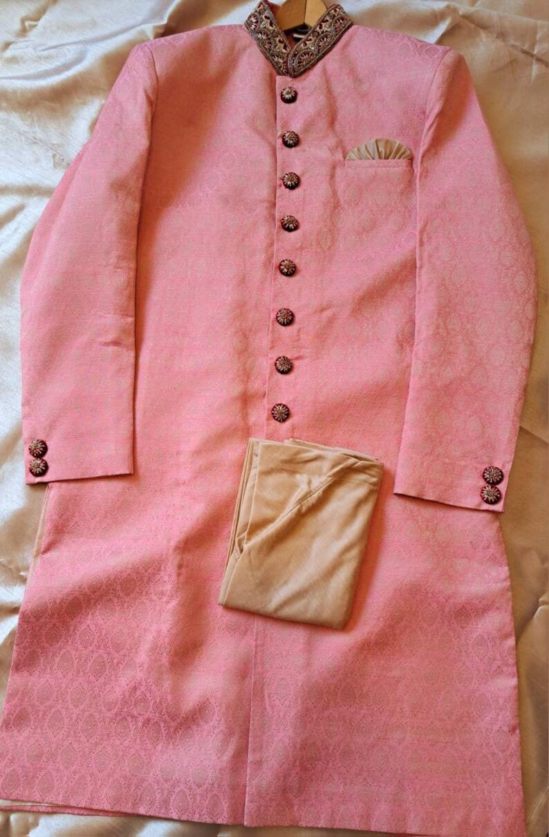 Puede incluir: Una t&uacute;nica rosa claro de manga larga con cuello mandar&iacute;n y botones decorativos. La t&uacute;nica tiene un sutil estampado floral y se combina con pantalones beige. El cuello y los pu&ntilde;os presentan bordados ornamentados.
