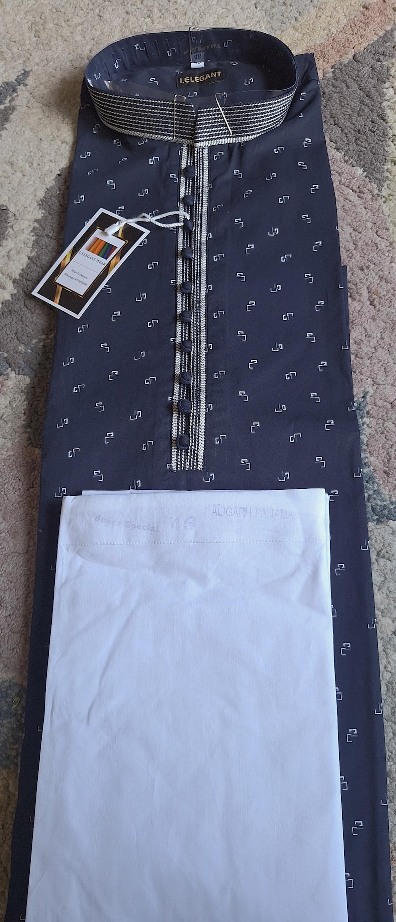 May include: Navy blue kurta pajama set. The kurta features a small white geometric pattern, a button-down front, and a decorative collar. The set includes a matching white pajama. A tag with the brand name is attached.