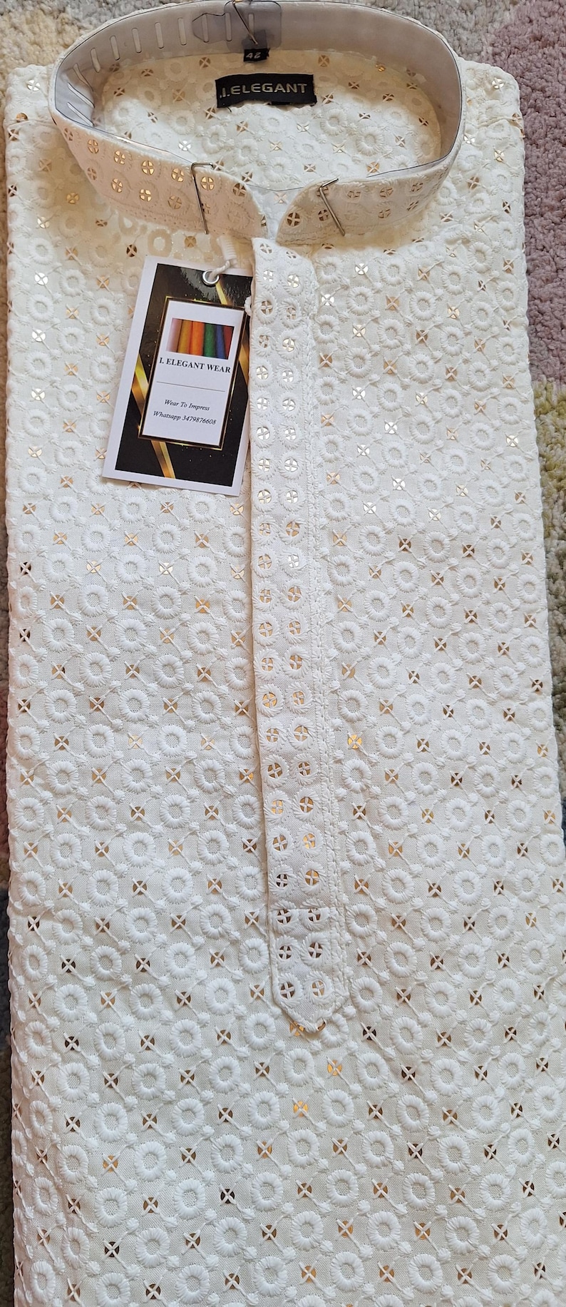 Puede incluir: Camisa blanca para hombre con un patr&oacute;n bordado en dorado. La camisa tiene un cuello mandar&iacute;n y cierre de bot&oacute;n. La etiqueta de la camisa dice "J. ELEGANT" e "I. ELEGANT WEAR".