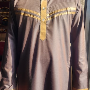 May include: A brown long-sleeved shirt with a gold trim around the collar and cuffs. The shirt has a button-up closure and a gold trim design on the front.