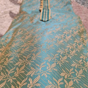May include: A teal-colored garment with a gold floral pattern. The fabric has a subtle sheen. The neckline is accented with a gold and teal striped design. The garment appears to be a traditional style.