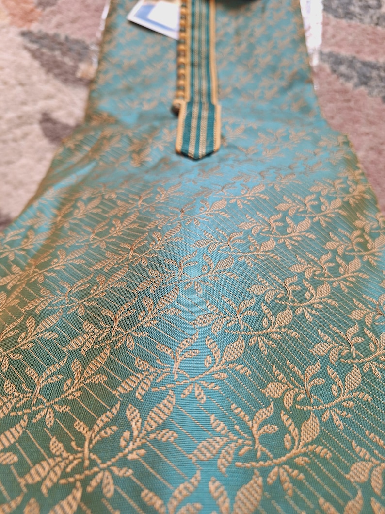 May include: A teal-colored garment with a gold floral pattern. The fabric has a subtle sheen. The neckline is accented with a gold and teal striped design. The garment appears to be a traditional style.