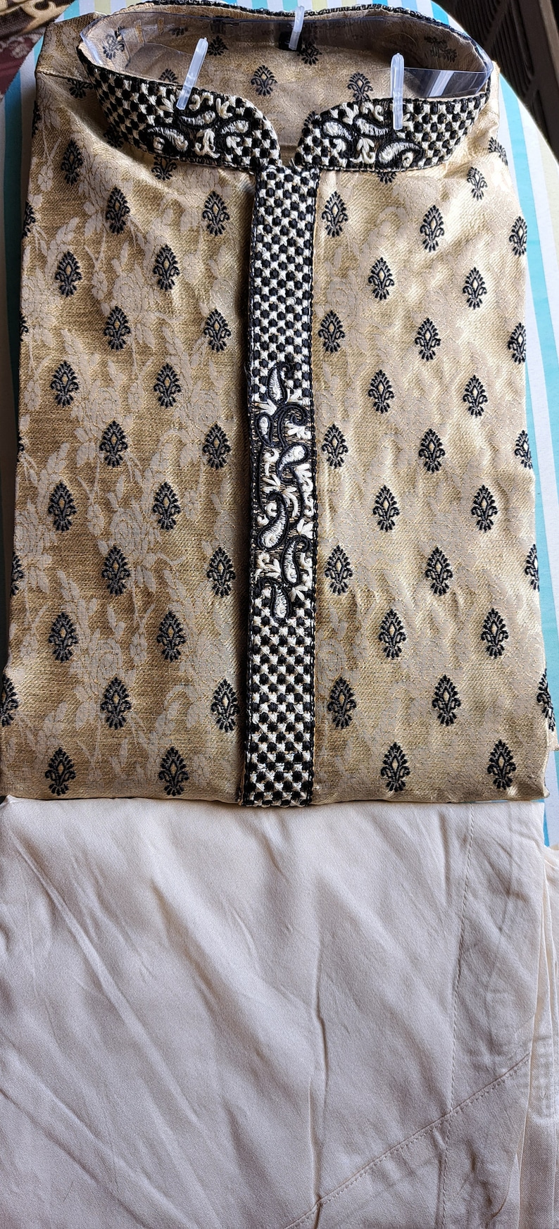 Op de afbeelding: Een gouden kurta met zwarte borduursels en een witte kraag. De kurta is gevouwen en gelegd op een witte stof.