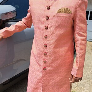 Puede incluir: Chaqueta de manga larga de color melocot&oacute;n claro con un patr&oacute;n floral detallado. La chaqueta presenta botones decorativos, un pa&ntilde;uelo de bolsillo dorado y un cuello detallado. La chaqueta forma parte de un atuendo formal tradicional.