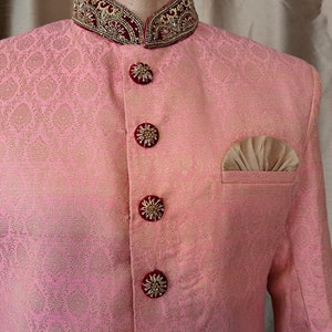 Puede incluir: Chaqueta rosa de brocado con estampado floral dorado. La chaqueta presenta botones decorativos con detalles dorados y burdeos, un pa&ntilde;uelo de bolsillo y un cuello adornado con bordados dorados y burdeos. Ropa formal.