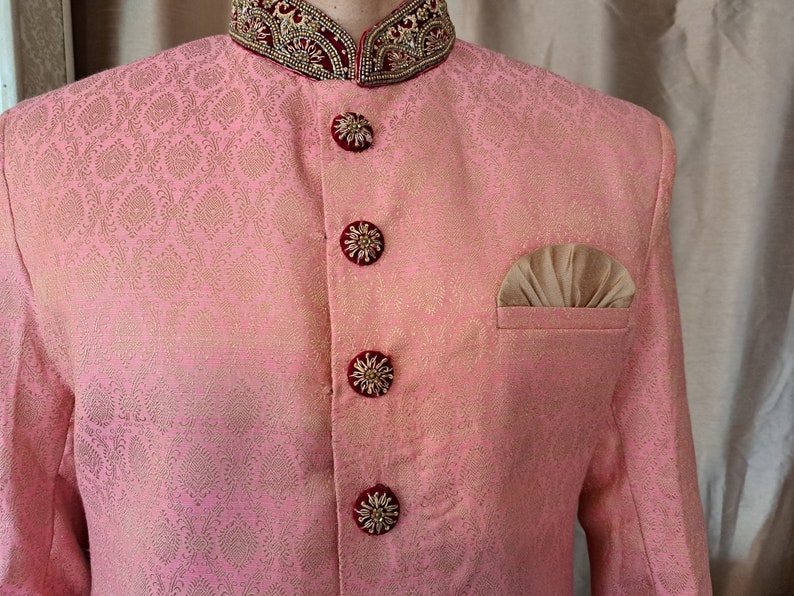 Puede incluir: Chaqueta rosa de brocado con estampado floral dorado. La chaqueta presenta botones decorativos con detalles dorados y burdeos, un pa&ntilde;uelo de bolsillo y un cuello adornado con bordados dorados y burdeos. Ropa formal.